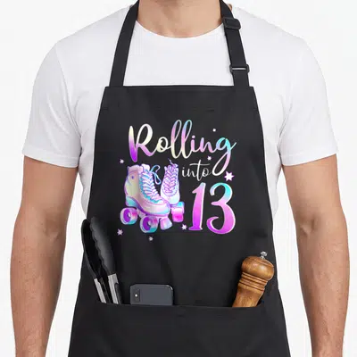 Rolling 13th birthday apron - Rolling into 13 text with roller skates and stars in purple blue yellow white — shown on a black cotton canvas apron against a white background
