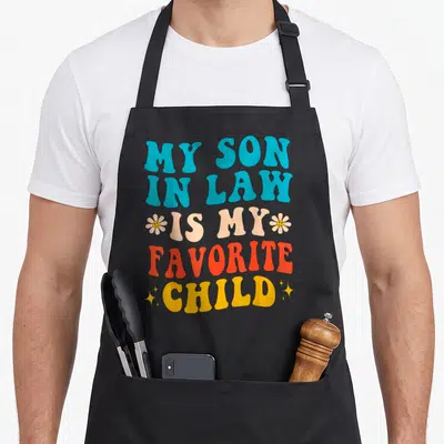 Retro law mother son aprons - Retro text MY SON IN LAW IS MY FAVORITE CHILD with smiley flowers and stars in blue peach — shown on a black cotton canvas apron against a white background
