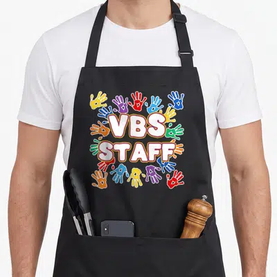 Vacation colorful aprons - VBS STAFF text surrounded by circular cluster of red, blue, yellow, green, purple — shown on a black cotton canvas apron against a white background