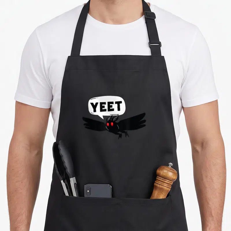 Mothman halloween birthday apron - Cartoon black moth with red eyes and white speech bubble reading yeet — shown on a black cotton canvas apron against a white background