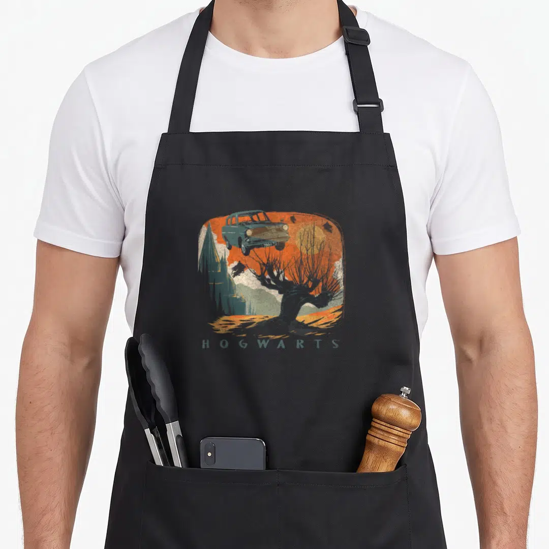 Whomping distressed apron - Retro flying car above tree and castle with HOGWARTS text in orange and grey-green — shown on a black cotton canvas apron against a white background