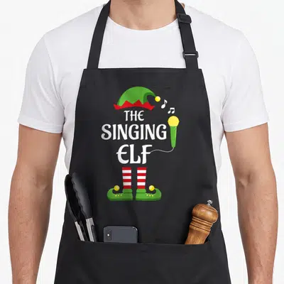 Singing family christmas elf apron - THE SINGING ELF text between green hat and red white striped legs with microphone — shown on a black cotton canvas apron against a white background