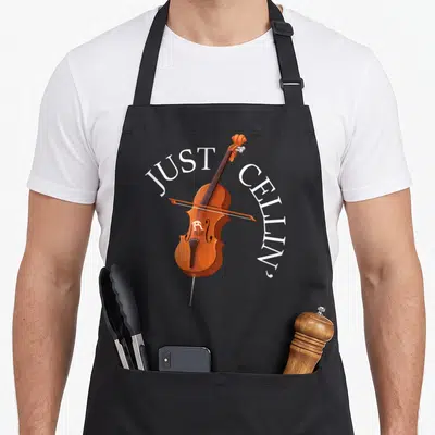 Player cello apron - Brown cartoon cello with white arched text JUST CELLIN' on gray background — shown on a black cotton canvas apron against a white background