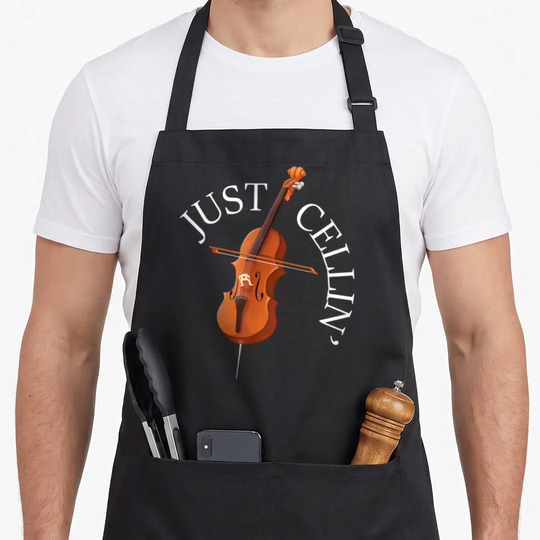 Player cello apron - Brown cartoon cello with white arched text JUST CELLIN' on gray background — shown on a black cotton canvas apron against a white background