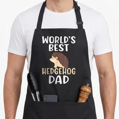 Retro hedgehog dad apron - Cartoon hedgehog flanked by WORLD'S BEST HEDGEHOG DAD text in white and beige — shown on a black cotton canvas apron against a white background