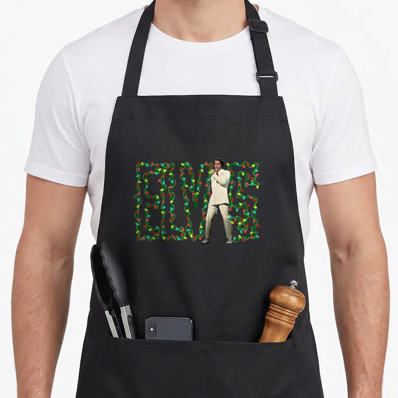 Elvis christmas lights apron - Photo of Elvis Presley in white suit standing before green ELVIS letters wrapped in red — shown on a black cotton canvas apron against a white background