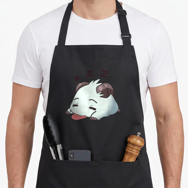 Tired master apron - White horned creature lying down with pink tongue and three dark brown Z letters floating above — shown on a black cotton canvas apron against a white background