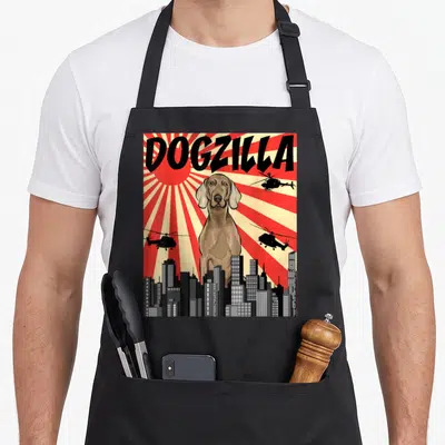 Japanese owner apron - DOGZILLA text above giant brown dog, three helicopters, city skyline, red sunburst — shown on a black cotton canvas apron against a white background