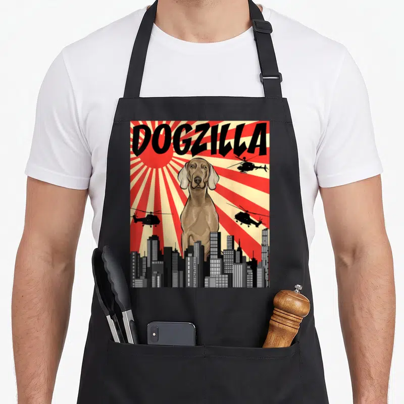 Japanese owner apron - DOGZILLA text above giant brown dog, three helicopters, city skyline, red sunburst — shown on a black cotton canvas apron against a white background