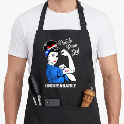 Puerto heritage aprons - Cartoon woman flexing arm with flag headband and UNBREAKABLE text — shown on a black cotton canvas apron against a white background