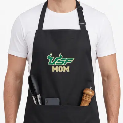 Bull florida apron - USF logo with bull horns above MOM text in green and gold typography — shown on a black cotton canvas apron against a white background