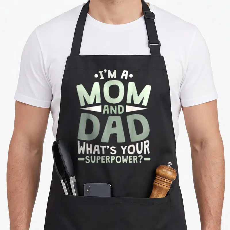 Superpower single mom apron - Stacked text I'm a Mom and Dad in white and green on black — shown on a black cotton canvas apron against a white background