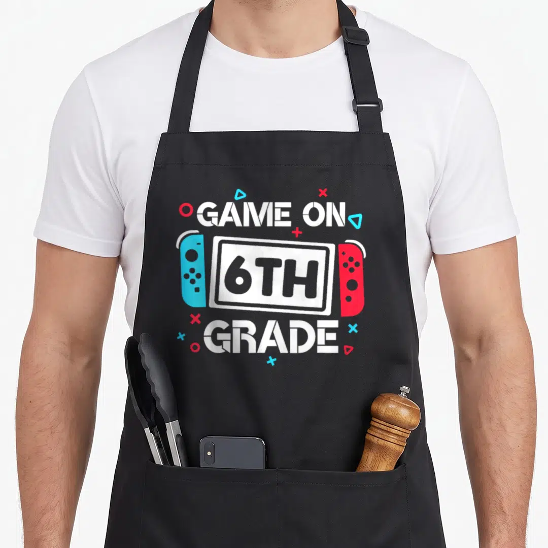 Game teacher apron - Game console with GAME ON 6TH GRADE text and scattered blue red symbols — shown on a black cotton canvas apron against a white background