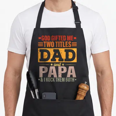 God father day apron - Stacked text design reading Dad and Papa in orange, pink, and olive distressed fonts — shown on a black cotton canvas apron against a white background