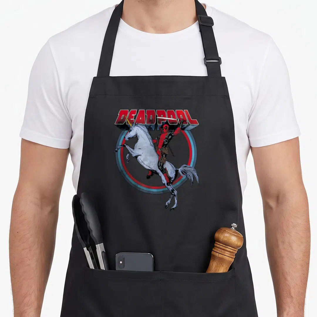 Merc rainbow apron - Deadpool riding a rearing white unicorn with a sword inside a red and blue circle — shown on a black cotton canvas apron against a white background