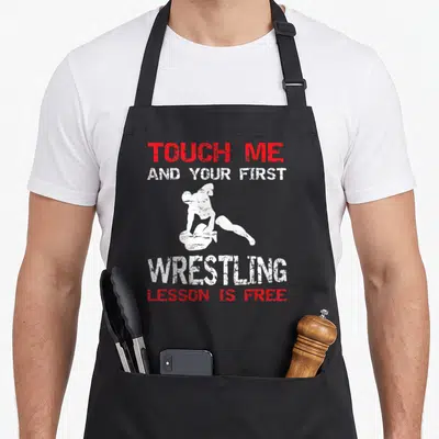 Lesson wrestling apron - Red and white distressed text touching wrestler silhouette pinning opponent — shown on a black cotton canvas apron against a white background