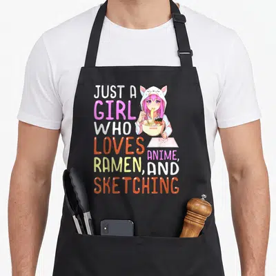 Ramen sketching anime apron - Pink hair anime girl in cat hoodie eating ramen and holding sketchbook with stacked — shown on a black cotton canvas apron against a white background