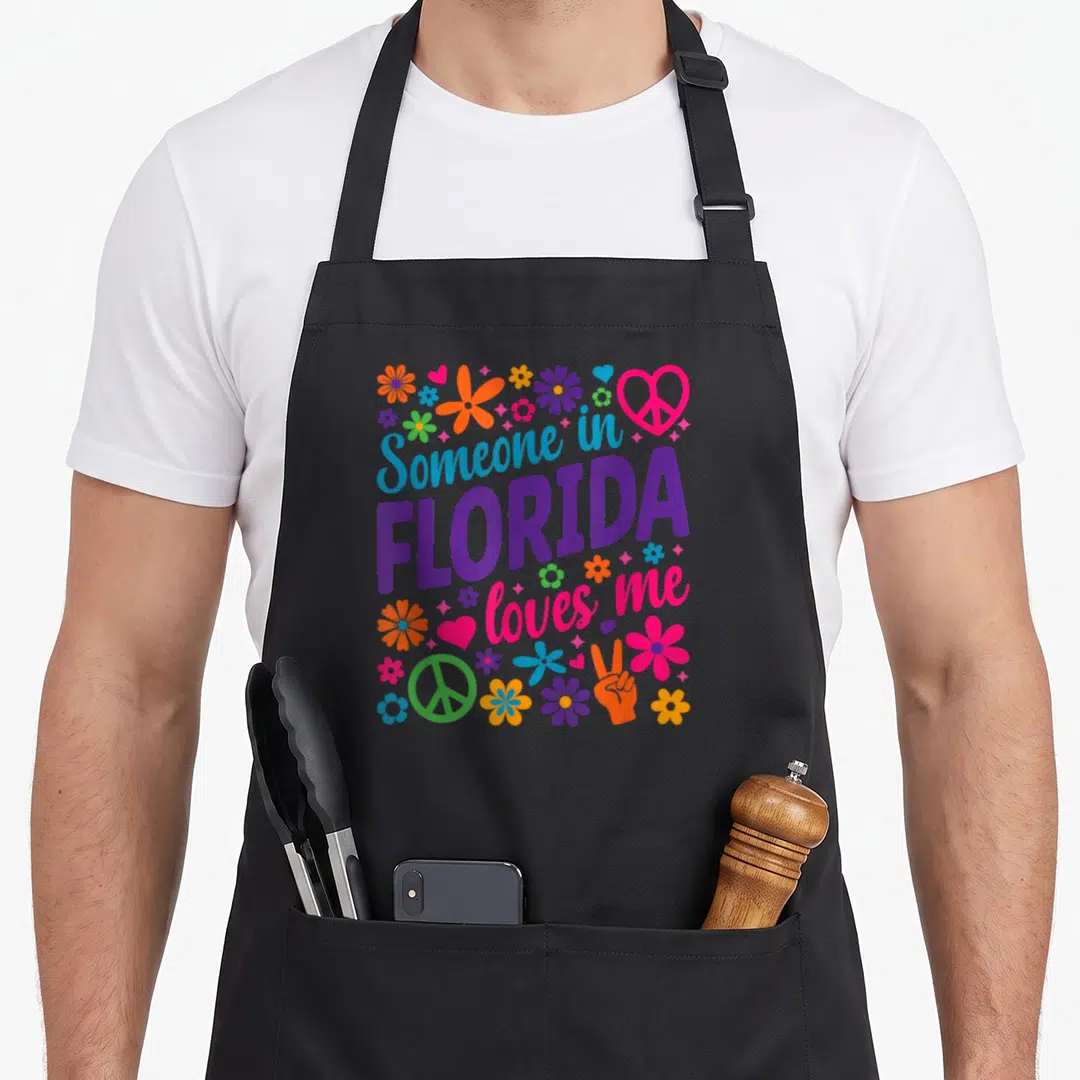 Someone florida apron - Stacked colorful text surrounded by cartoon flowers, hearts, and peace signs in purple pink — shown on a black cotton canvas apron against a white background
