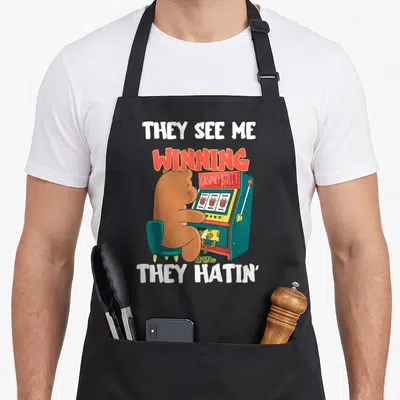 Machine casino aprons - Brown bear playing teal slot machine with text THEY SEE ME WINNING THEY HATIN' — shown on a black cotton canvas apron against a white background