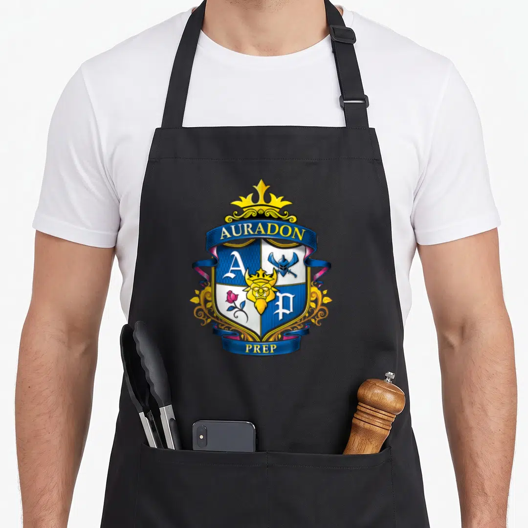 Auradon novelty apron - Auradon Prep crest with blue ribbons, gold crown, shield quadrants, rose, helmet, and lion head — shown on a black cotton canvas apron against a white background