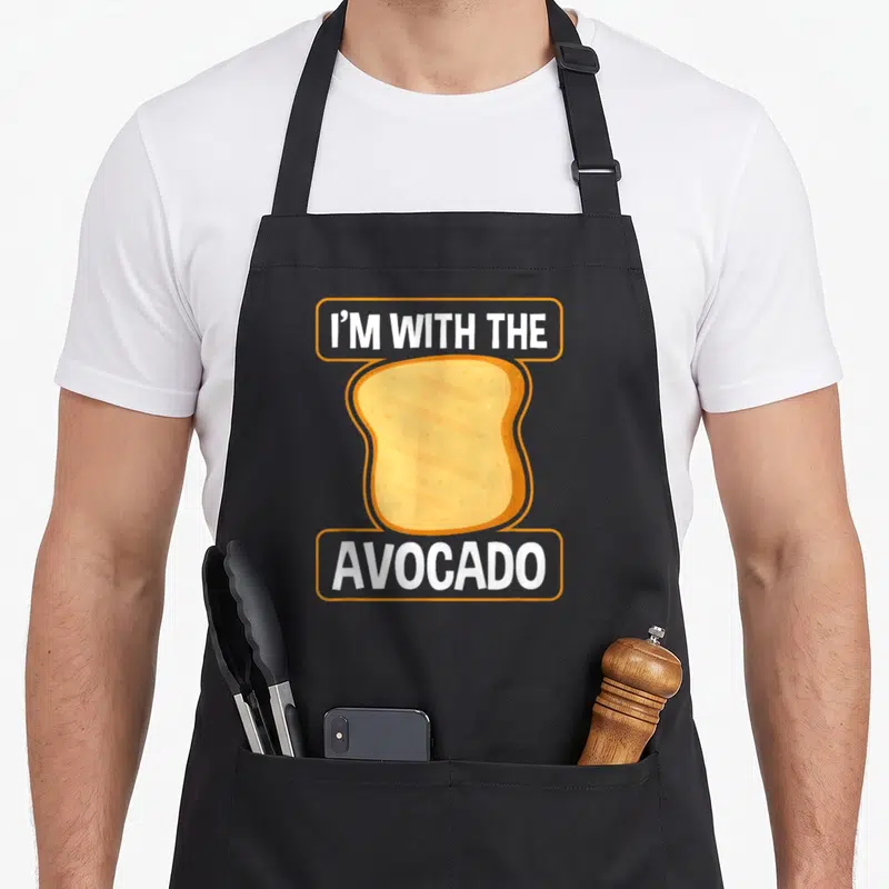 Avocado halloween toast apron - Cartoon toast between two orange banners with white text reading I M WITH THE AVOCADO — shown on a black cotton canvas apron against a white background