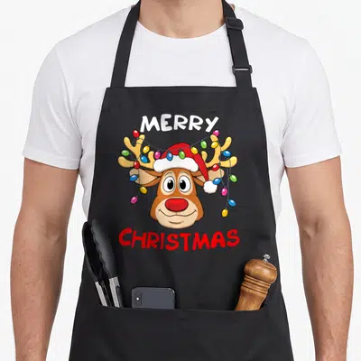 Reindeer merry christmas apron - Cartoon reindeer head with Santa hat and colorful lights, Merry Christmas text — shown on a black cotton canvas apron against a white background