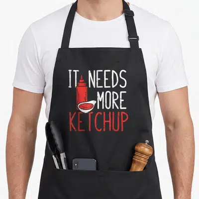 Funny novelty ketchup apron - Distressed red ketchup bottle and bowl with IT NEEDS MORE KETCHUP text — shown on a black cotton canvas apron against a white background