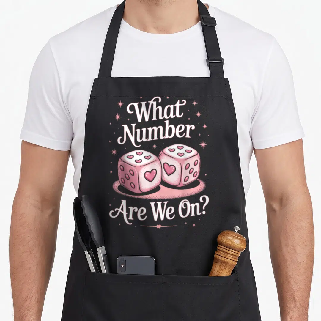 Number game aprons - Two pink dice with heart pips and white text What Number Are We On surrounded by stars — shown on a black cotton canvas apron against a white background