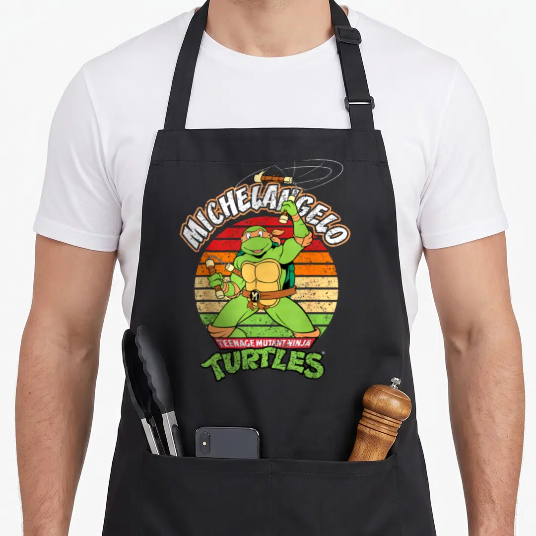 Michelangelo ready teenage cooking aprons - Green cartoon turtle holding nunchaku in front of a striped red orange — shown on a black cotton canvas apron against a white background