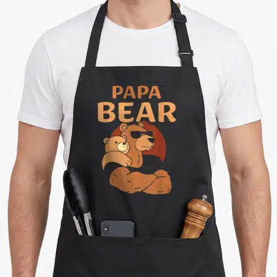 Papa daddy apron - Orange cartoon Papa Bear holding cub with stacked text — shown on a black cotton canvas apron against a white background