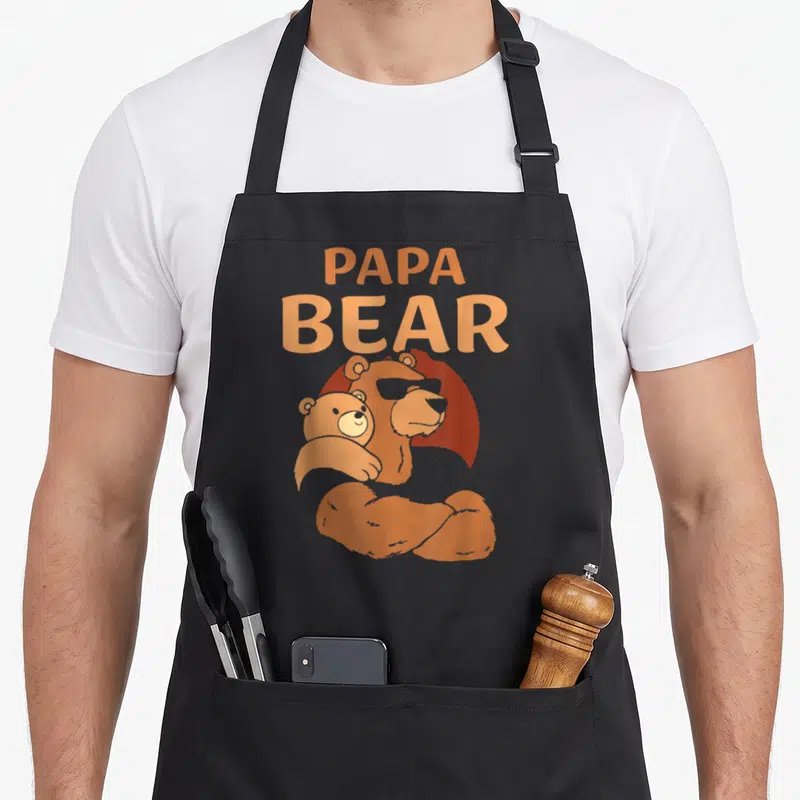 Papa daddy apron - Orange cartoon Papa Bear holding cub with stacked text — shown on a black cotton canvas apron against a white background