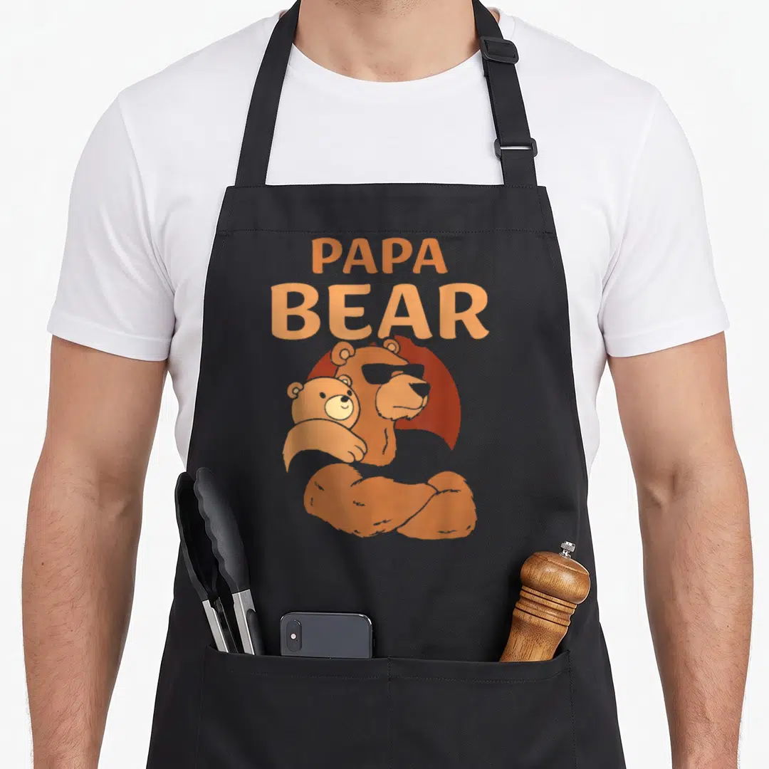 Papa daddy apron - Orange cartoon Papa Bear holding cub with stacked text — shown on a black cotton canvas apron against a white background