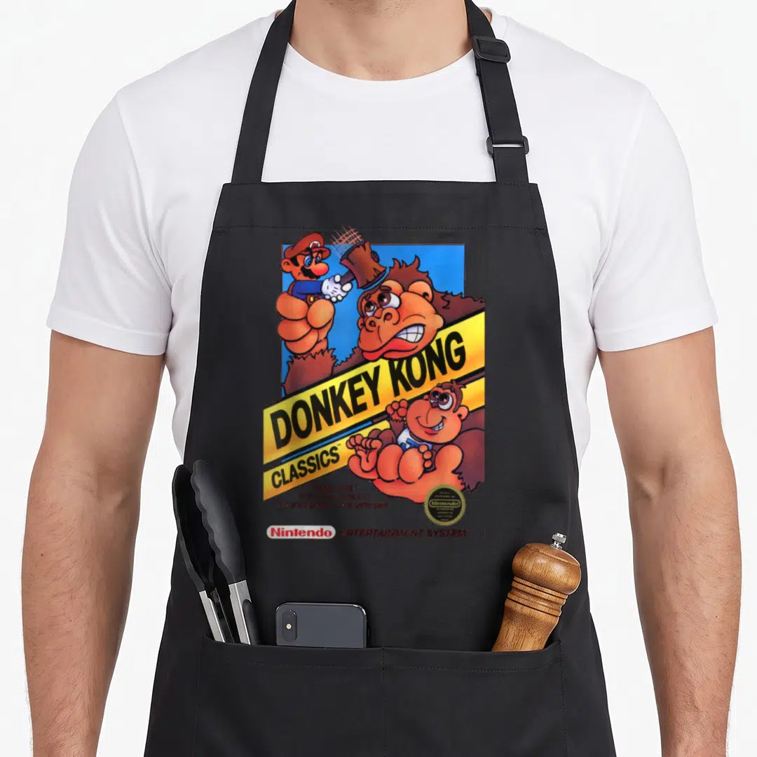 Hammer donkey apron - Donkey Kong Classics cover art with Mario holding a hammer and yellow banner on blue background — shown on a black cotton canvas apron against a white background