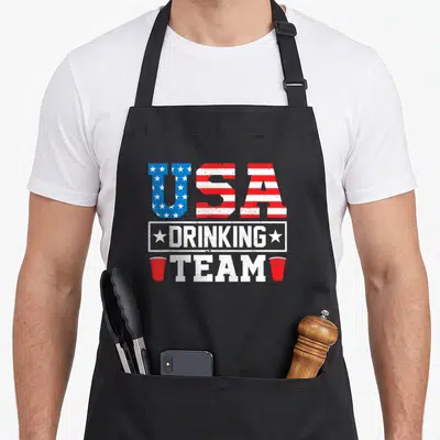 Retro drinking beer apron - Distressed USA DRINKING TEAM text with flag fills and two red cups — shown on a black cotton canvas apron against a white background