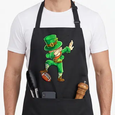 Football st patrick day aprons - Cartoon leprechaun dabbing on football in green and brown — shown on a black cotton canvas apron against a white background