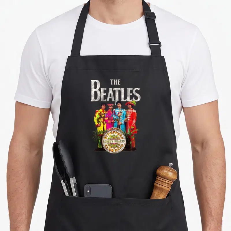 Sgt lonely pepper apron - The Beatles four members in colorful uniforms around a drum with album title text and plants — shown on a black cotton canvas apron against a white background