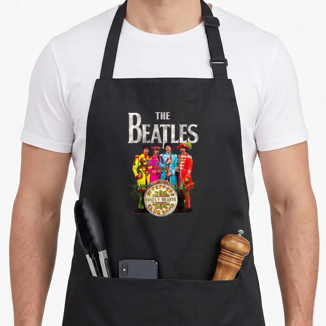 Sgt lonely pepper apron - The Beatles four members in colorful uniforms around a drum with album title text and plants — shown on a black cotton canvas apron against a white background
