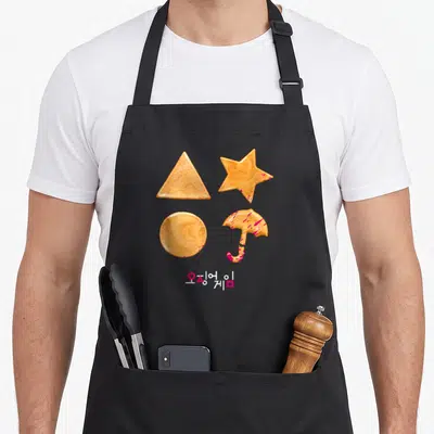 Sugar cookies apron - Four orange-brown dalgona candies with pink splatters and Korean text below — shown on a black cotton canvas apron against a white background