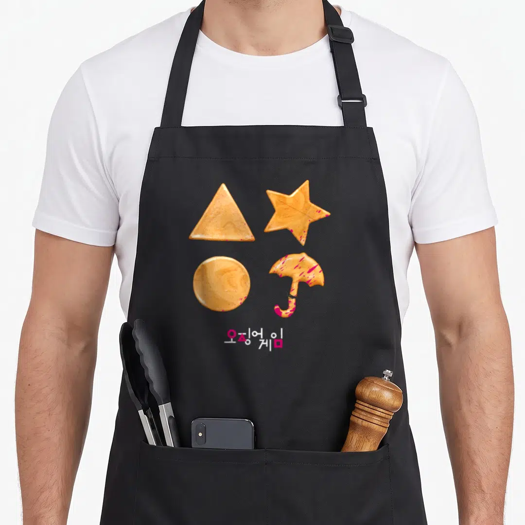 Sugar cookies apron - Four orange-brown dalgona candies with pink splatters and Korean text below — shown on a black cotton canvas apron against a white background