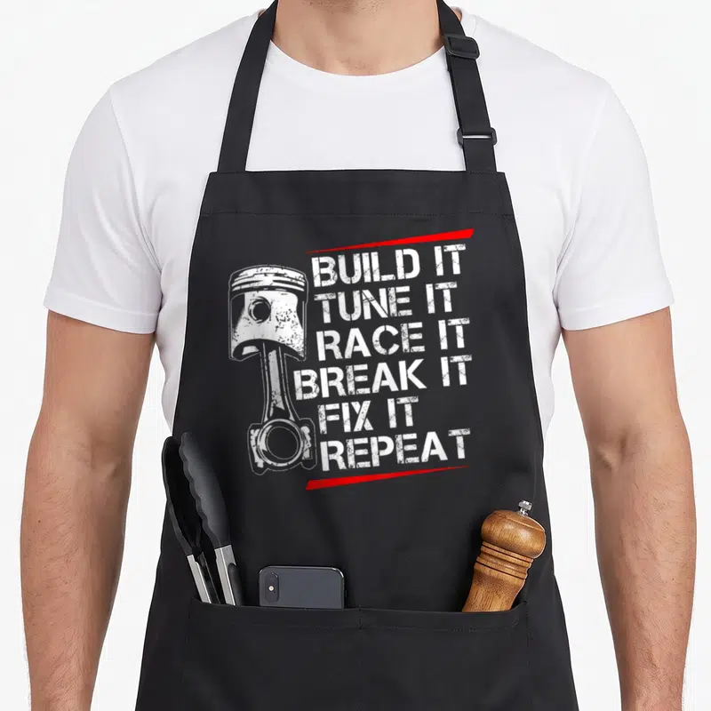Step car apron - White piston and six distressed lines of text framed by two red stripes — shown on a black cotton canvas apron against a white background