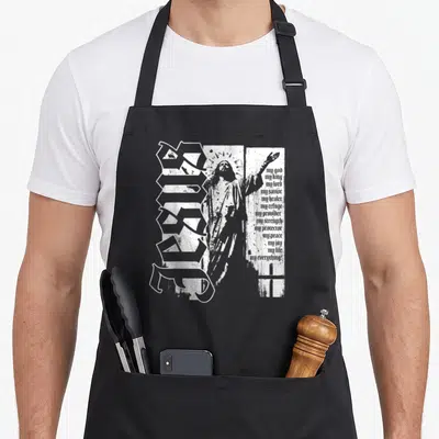 Christ king of the grill apron - Distressed white jesus figure with halo and vertical jesus text alongside stacked — shown on a black cotton canvas apron against a white background