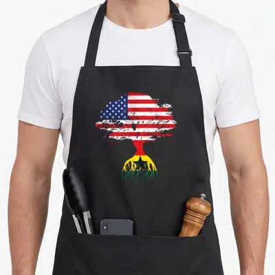 Ghanaian american flag apron - Tree with US flag canopy and Ghana flag roots in red white blue yellow green — shown on a black cotton canvas apron against a white background