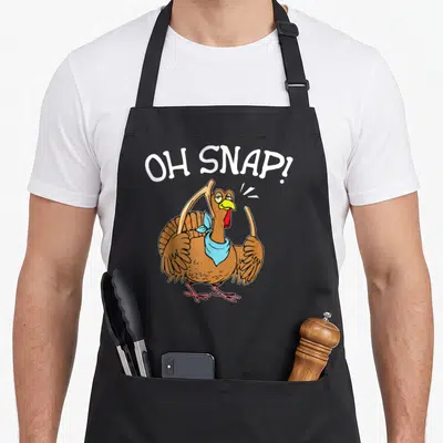 Broking thanksgiving day aprons - Cartoon turkey holding broken bones under arched OH SNAP text in brown and blue — shown on a black cotton canvas apron against a white background