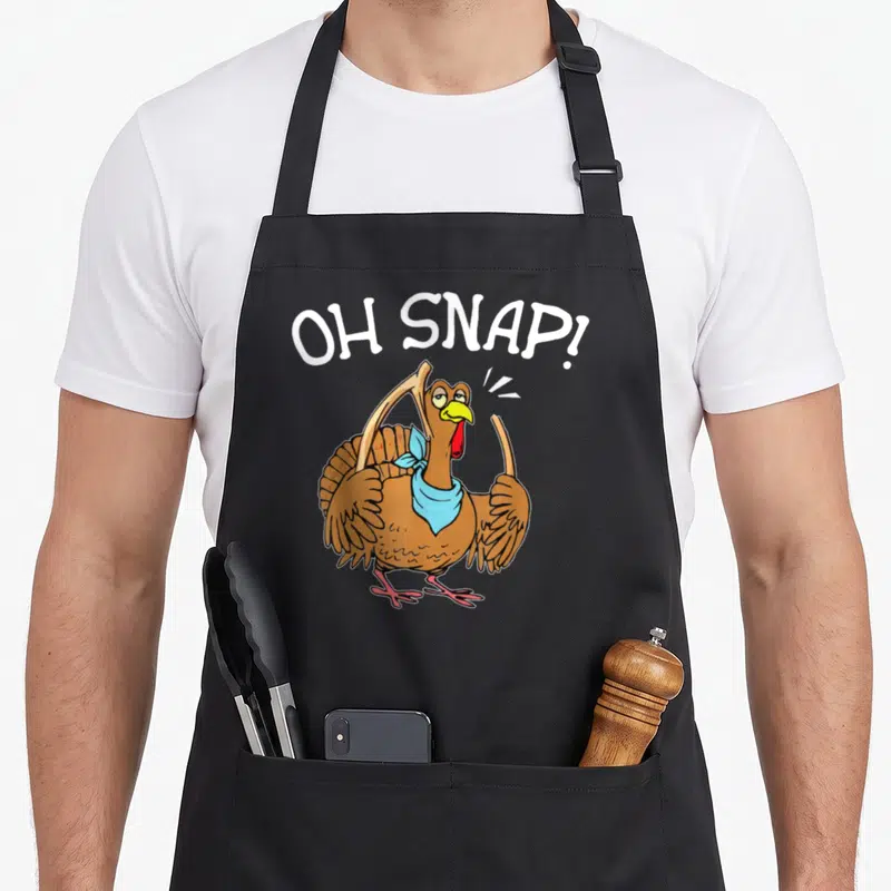 Broking thanksgiving day aprons - Cartoon turkey holding broken bones under arched OH SNAP text in brown and blue — shown on a black cotton canvas apron against a white background