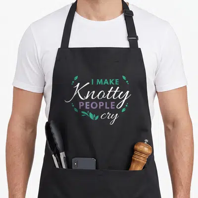 Cry massage therapist apron - Stacked text I Make Knotty People Cry with teal leaves and purple accents — shown on a black cotton canvas apron against a white background
