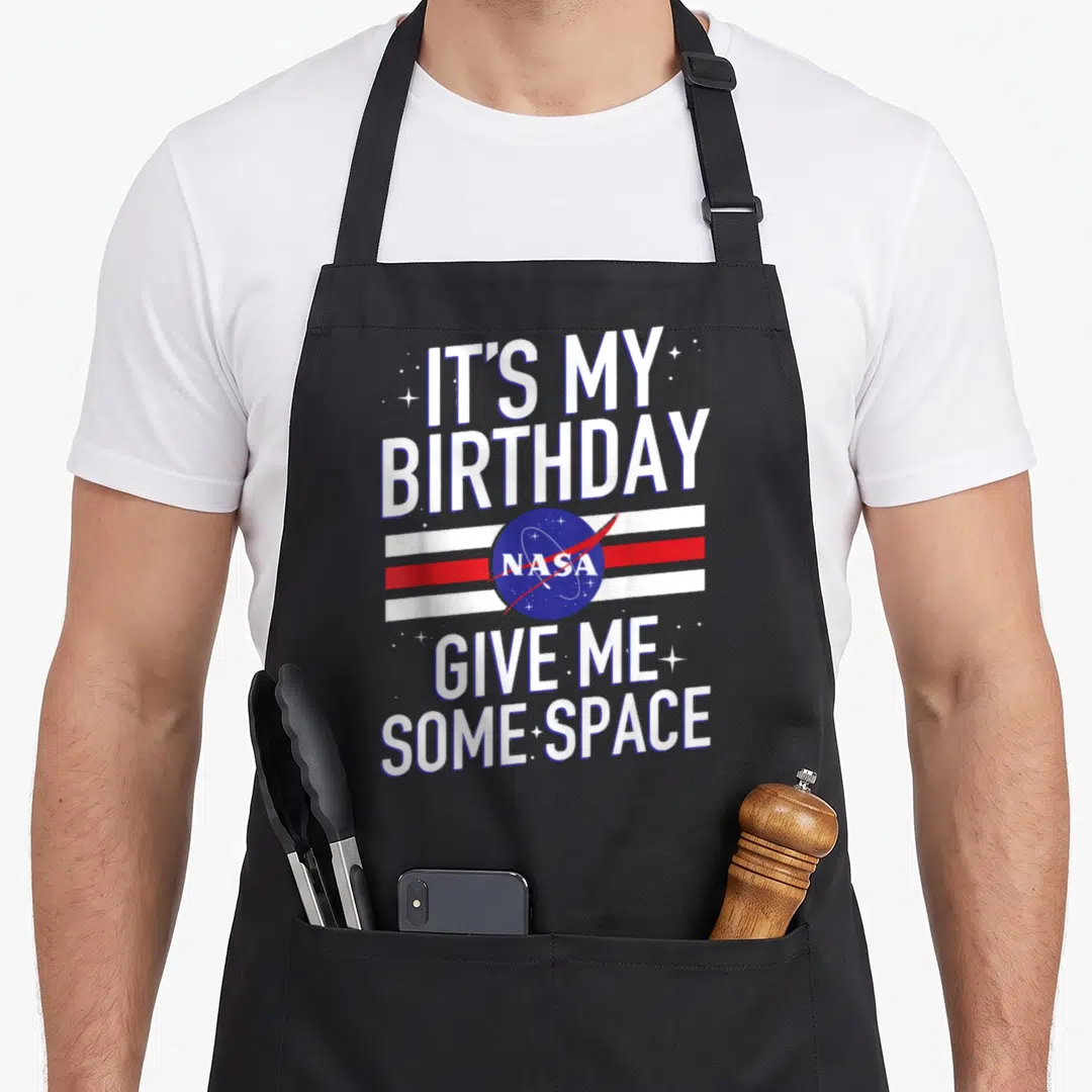 Birthday nasa apron - IT'S MY BIRTHDAY NASA GIVE ME SOME SPACE text with logo and stripes — shown on a black cotton canvas apron against a white background