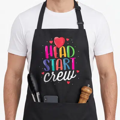 Start preschool teacher apron - stacked HEAD START crew text in pink yellow green blue red and white with hearts — shown on a black cotton canvas apron against a white background