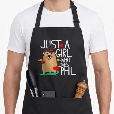 Punxsy holiday apron - Cartoon brown beaver in top hat with white text and red hearts on green grass — shown on a black cotton canvas apron against a white background