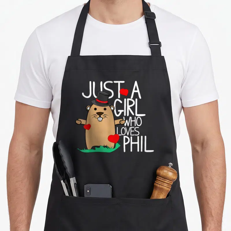 Punxsy holiday apron - Cartoon brown beaver in top hat with white text and red hearts on green grass — shown on a black cotton canvas apron against a white background