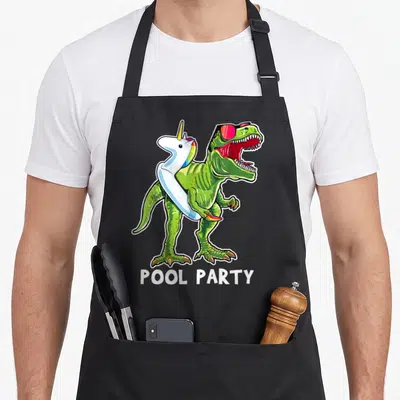 Pool dinosaur apron - Green T-Rex with red sunglasses and white unicorn floatie, POOL PARTY text below — shown on a black cotton canvas apron against a white background